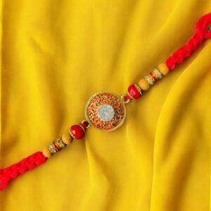 Traditional Rakhi with round Kundan-style centerpiece on red thread, displayed on a yellow fabric background