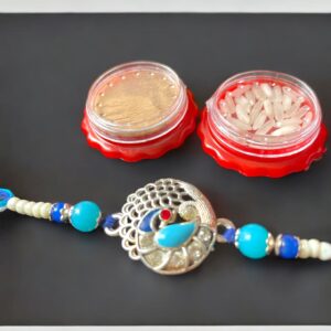 Silver-toned Peacock Rakhi with blue and white beads, accompanied by red roli and chawal containers on a black background